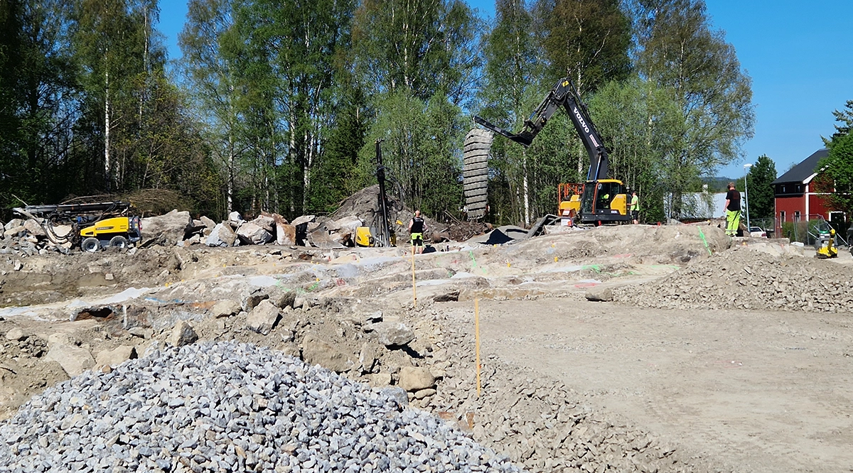 Bilden visar en byggarbetsplats med stenar och grus samt en grävskopa som jobbar på. Två arbetsklädda personer syns också.