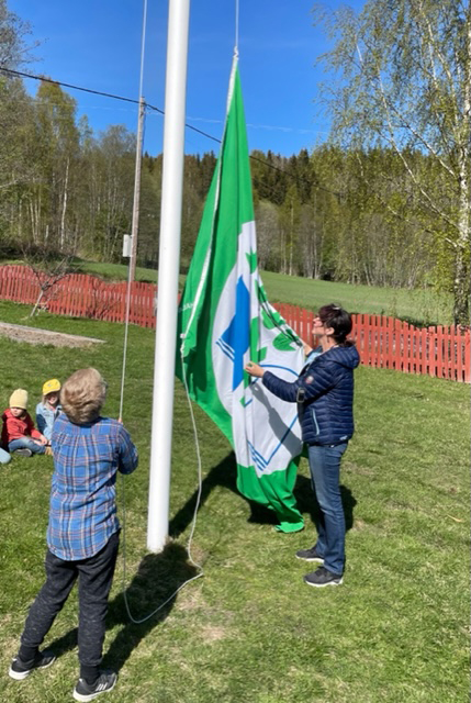 En vuxen och ett barn hissar en grön flagga vid en flaggstång.