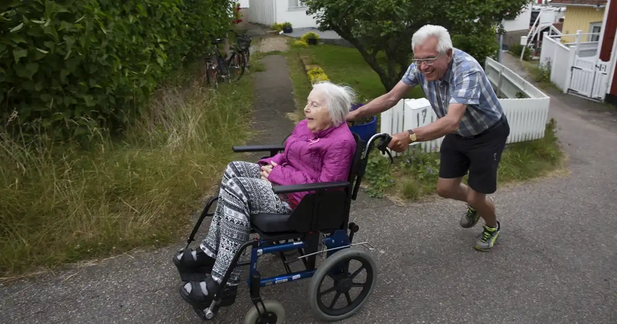 En gråhårig man i sommarkläder drar än äldre kvinna i rullstol uppför en backe. Båda skrattar.