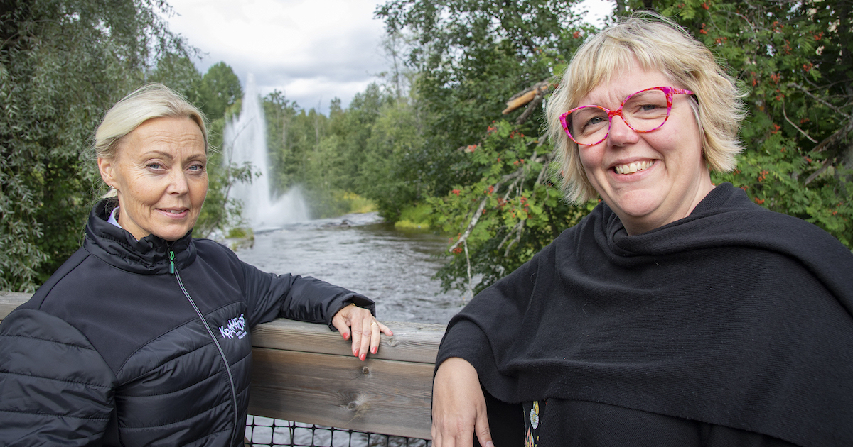 Två kvinnor på en bro framför en å med en fontän.
