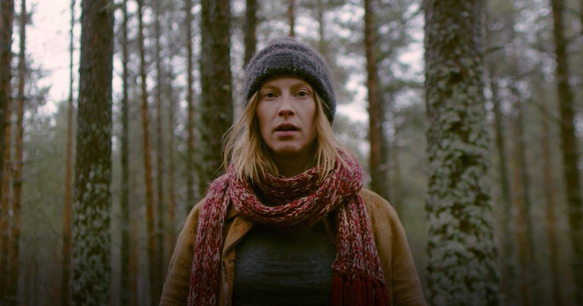 Kvinna står i skogen, mitt i bild och tittar rakt in i kameran. Synlig i halvprofil iklädd mössa halsduk jacka. 