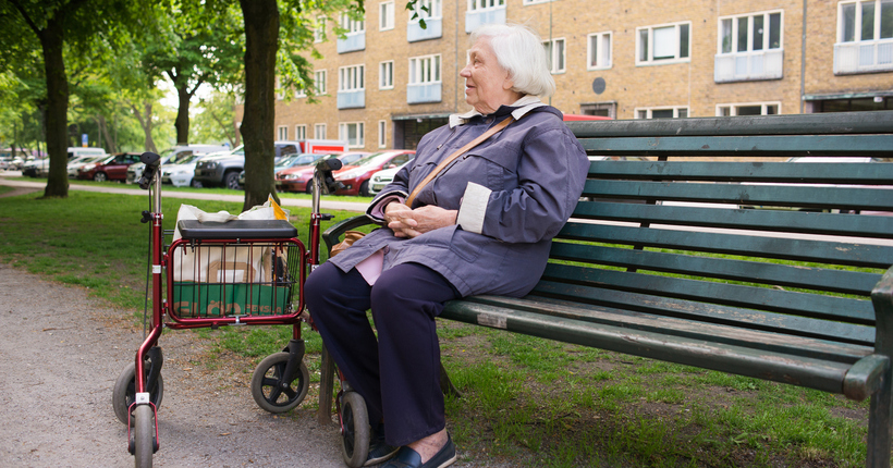 Äldre kvinna med rullator sitter på bänk.